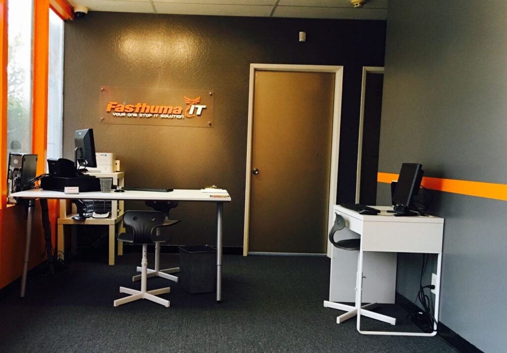 Inside the Fasthuma iT office in Fremont: two desks with computers and the Fasthuma iT logo sign on a gray wall